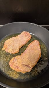 Zwei panierte Schnitzel braten in einer flachen Fettschicht in einer großen dunklen Pfanne, die Panade ist an den Rändern noch hell