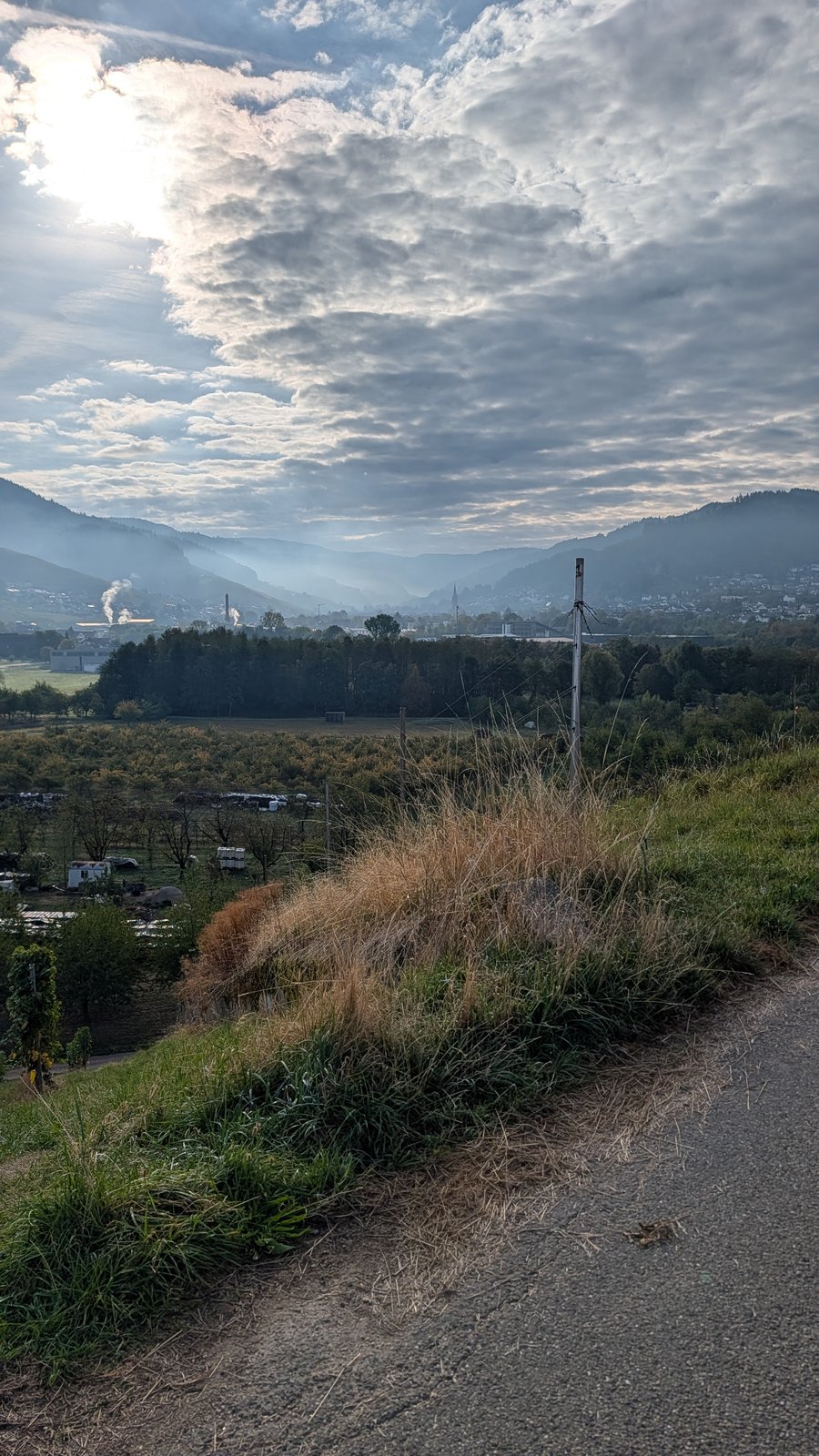 Schotterweg mit trockenem Gras und einem Holzpfosten an einem Hang, dahinter ein dunstiges Tal mit einer kleinen Stadt und bewaldeten Hügeln unter bewölktem Himmel