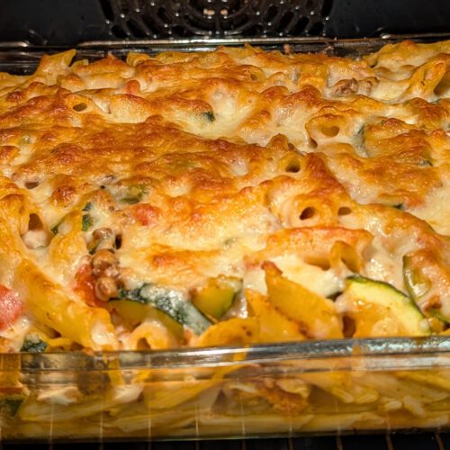 Fertig gebackener Penne-Auflauf in einer rechteckigen Glasform im Ofen, mit goldbrauner, blubbernder Käsehaube über Penne und Zucchini