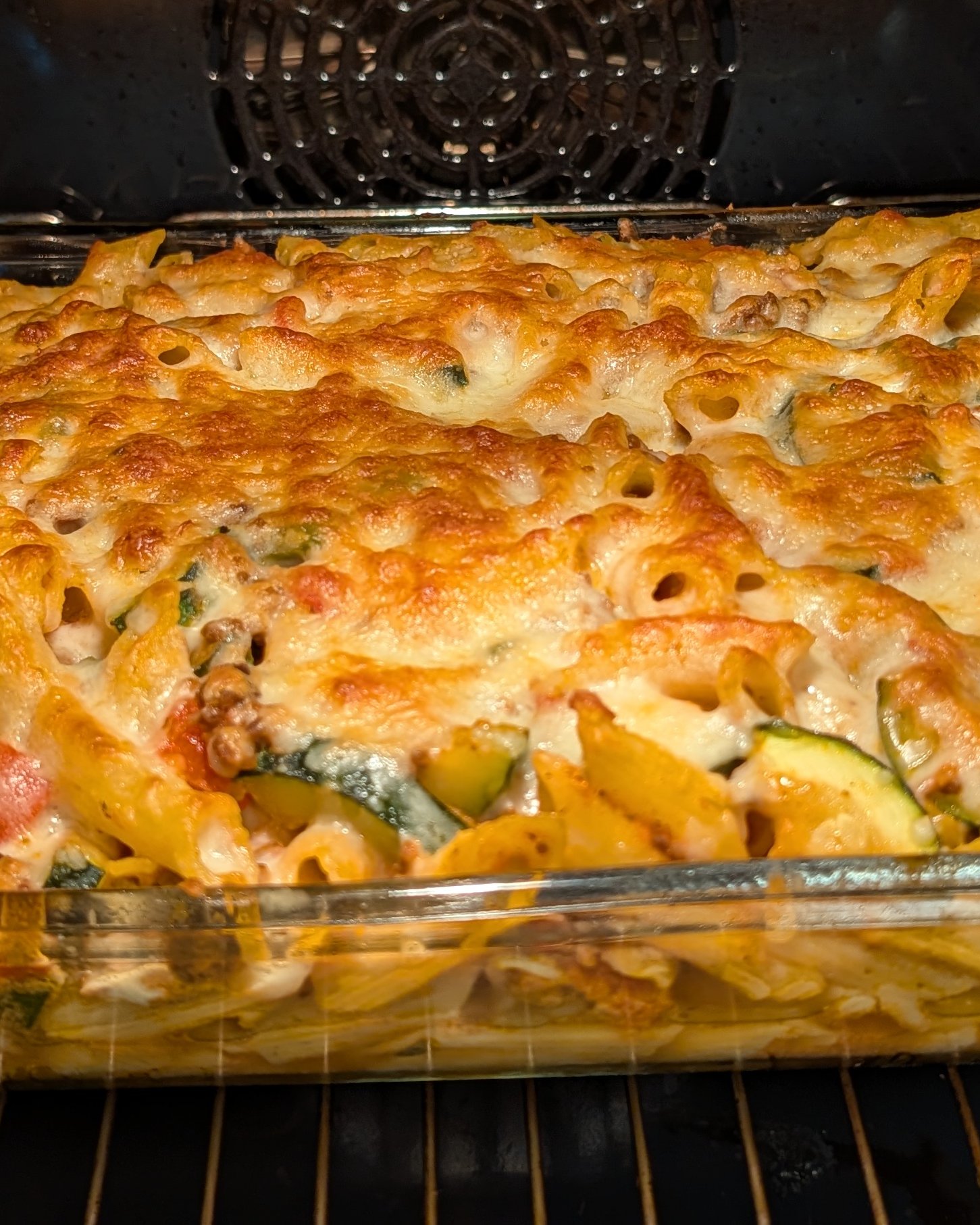Fertig gebackener Penne-Auflauf in einer rechteckigen Glasform im Ofen, mit goldbrauner, blubbernder Käsehaube über Penne und Zucchini