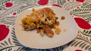 Eine Portion Penne-Auflauf auf einem weißen Teller mit brauner Käsekruste, geschmolzenen Käsefäden, Hacksoße und Zucchini, auf einem gemusterten Tischtuch mit roten Äpfeln