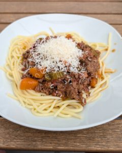 Teller mit Spaghetti, darauf Ragù alla Bolognese und ein Häufchen geriebener Käse, auf einem Holztisch