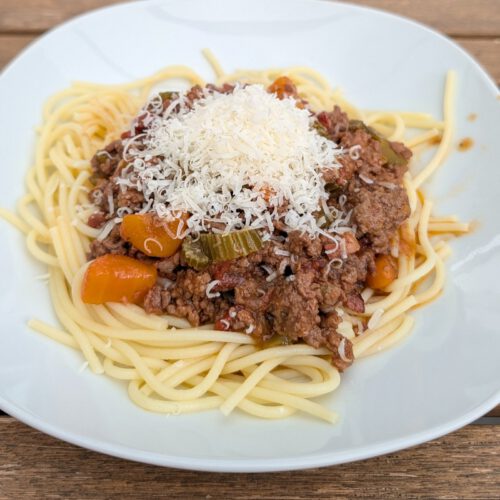 Teller mit Spaghetti, darauf Ragù alla Bolognese und ein Häufchen geriebener Käse, auf einem Holztisch