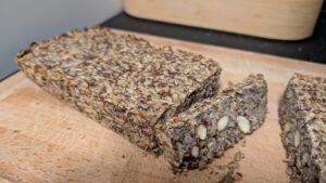 Gebackenes Saatenbrot mit flacher Oberseite und einer abgeschnittenen Scheibe, in der dichten Krume sind ganze Mandeln und Samen zu sehen
