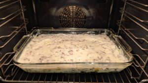 Ungebackene Moussaka in einer rechteckigen Glasform im Ofen, bedeckt mit einer hellen cremigen Soße, durch die Hackfleischkrümel schimmern.